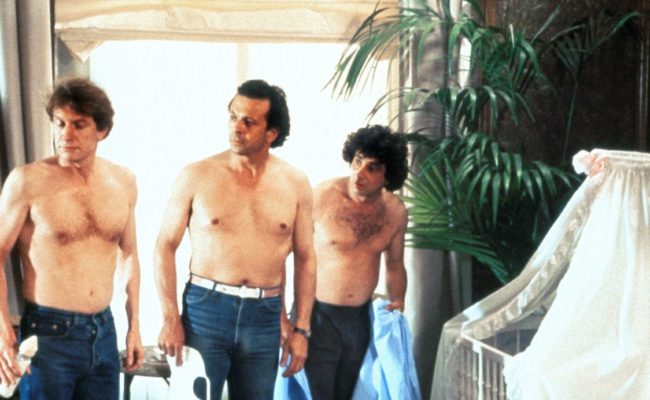 Scène du film 3 Hommes et un couffin. Trois hommes torse nu, en jean, debout dans un salon luxueux, regardent vers un berceau couvert de dentelle blanche. Chacun tient une chemise ou un accessoire de bébé, l'air surpris ou désorienté.