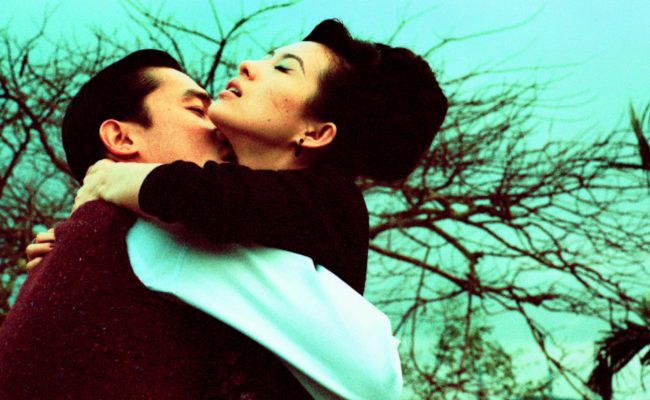 Un couple s’étreint passionnément sous un ciel turquoise, dans un décor extérieur aux arbres dénudés. L’homme embrasse la femme dans le cou, tandis qu’elle ferme les yeux, la tête renversée en arrière.