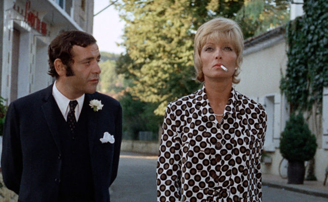 Un homme en costume sombre marche aux côtés d'une femme blonde en robe à pois, qui fume une cigarette, dans une rue calme bordée de fleurs et de bâtiments. En arrière-plan, une enseigne indique "Hôtel Restaurant".