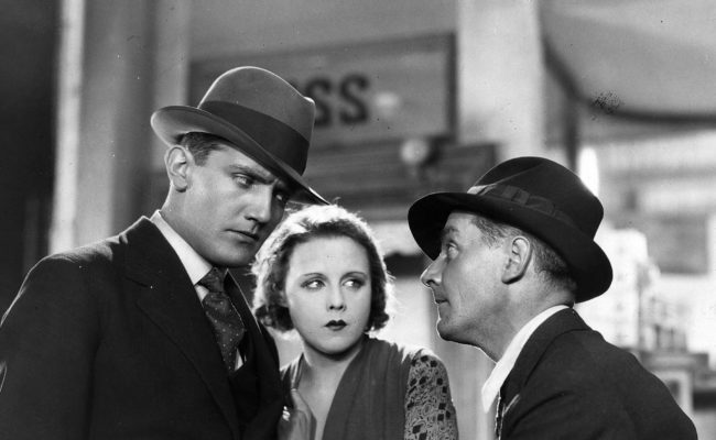 Photographie en noir et blanc issue du film Chantage d’Alfred Hitchcock. Trois personnages en costume des années 1920-1930 sont en pleine discussion tendue. Un homme au regard dur et méfiant fixe un autre homme, qui semble tenter de le raisonner en posant une main sur lui. Entre eux, une femme au maquillage marqué les observe avec inquiétude, posant une main sur le bras du premier.