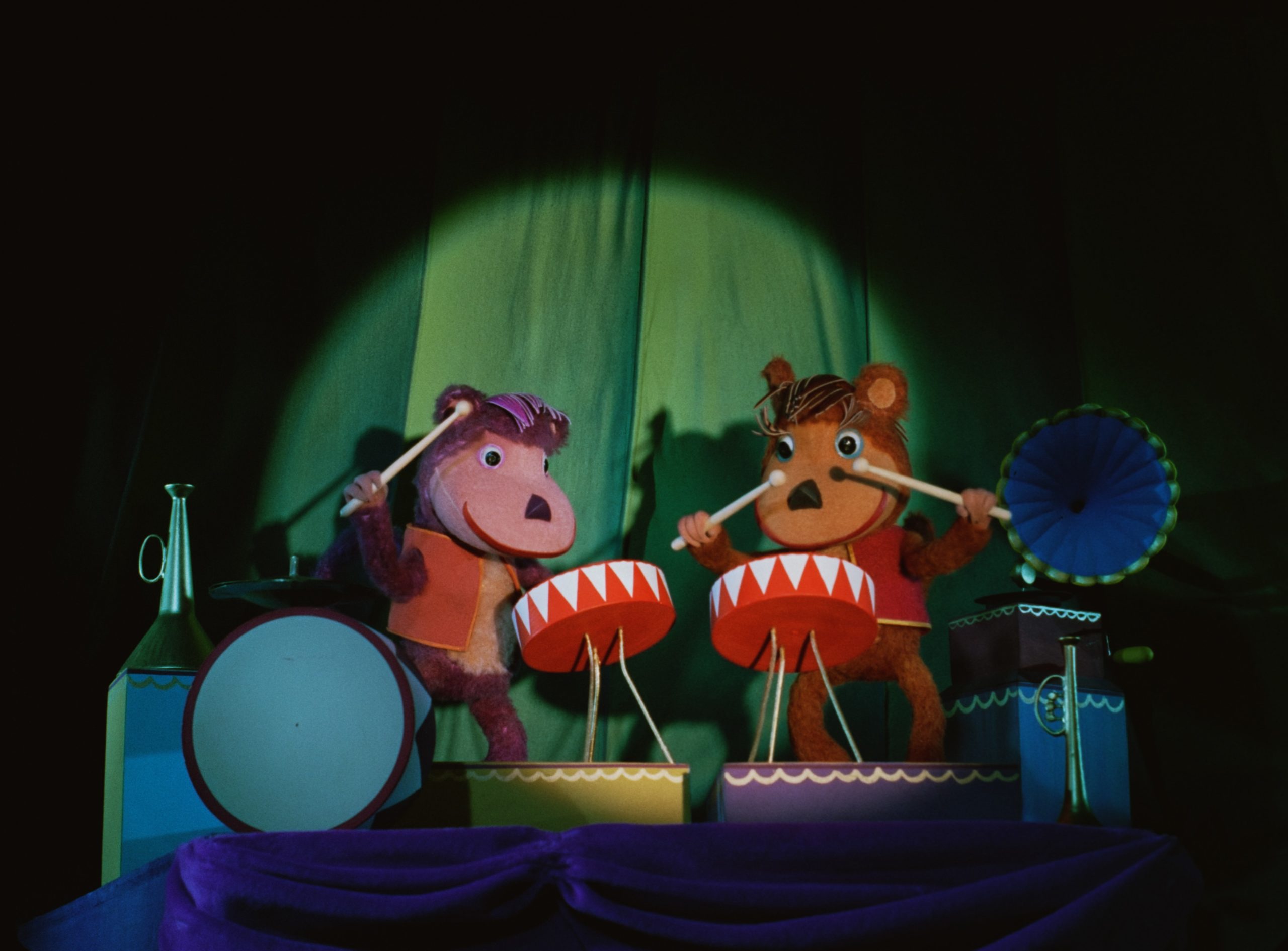 Deux marionnettes animales en peluche, l’une violette et l’autre orange, jouent du tambour sur une scène de cirque. Elles sont éclairées par un projecteur circulaire sur un rideau vert. Chaque personnage est debout devant un tambour rouge et blanc, tenant des baguettes. À droite, un vieux haut-parleur bleu est posé sur une boîte. Des éléments de décor colorés, comme un tambourin, complètent la scène. L’ambiance évoque un spectacle musical pour enfants en stop-motion.