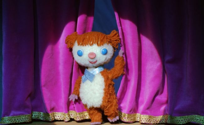 Une marionnette en peluche représentant un petit ourson orange et blanc, avec un nœud papillon bleu scintillant, écarte un rideau de velours rose vif bordé de doré. L’ourson, souriant, est éclairé par un projecteur bleu alors qu’il entre en scène, dans un décor évoquant un théâtre de marionnettes.