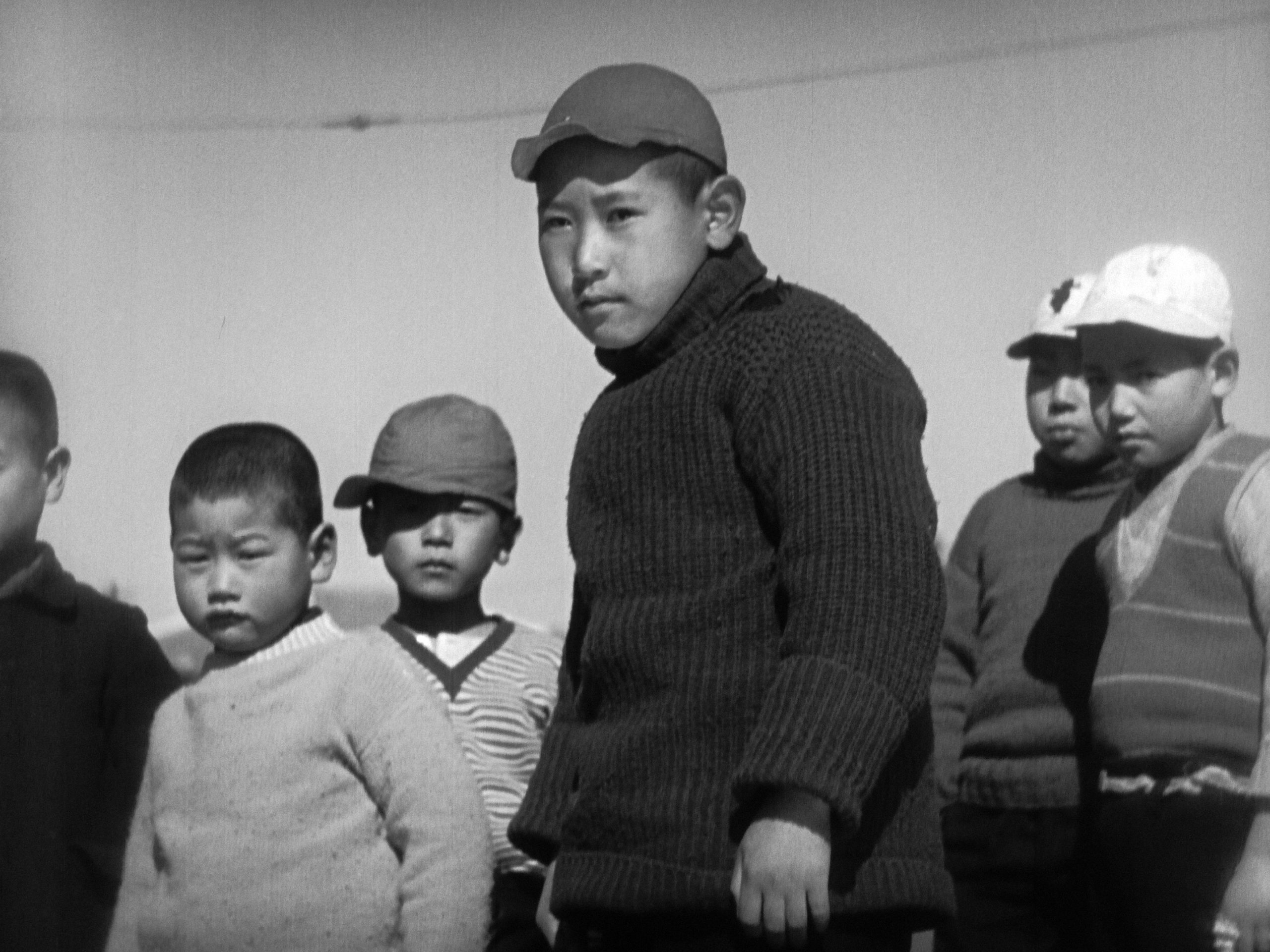 Photographie en noir et blanc d’un groupe de jeunes garçons japonais vêtus de pulls et de casquettes. Le garçon au centre, au premier plan, porte un pull foncé et une casquette claire, et regarde au loin avec sérieux. Les autres enfants, en arrière-plan, semblent attentifs ou songeurs, sous un ciel dégagé.