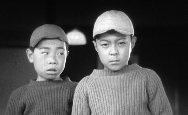 Image en noir et blanc de deux garçons japonais côte à côte, vêtus de pulls épais et de casquettes. Le garçon de droite regarde fixement l’objectif avec un air grave, tandis que celui de gauche tourne légèrement la tête vers lui avec une expression intriguée. L’arrière-plan est sombre, soulignant les visages et les expressions des enfants.