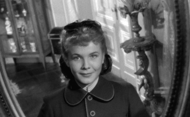 Reflet d’une jeune femme souriante dans un miroir ovale ancien, situé dans un intérieur bourgeois. Elle porte un manteau à col Claudine et boutons visibles, et fixe son propre reflet avec un sourire.