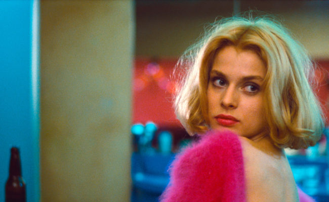 Image tirée du film Paris, Texas de Wim Wenders. Une femme blonde au carré parfait, maquillée avec soin, est assise de dos, légèrement tournée vers la caméra. Elle porte un pull rose fuchsia en mohair, tombant sur ses épaules nues. Elle regarde en arrière avec une expression à la fois distante et mélancolique. L’arrière-plan est un décor aux lumières bleutées et rougeâtres, suggérant un lieu intime. L’image est saturée de couleurs vives, soulignant l’atmosphère électrique et émotionnelle de la scène.