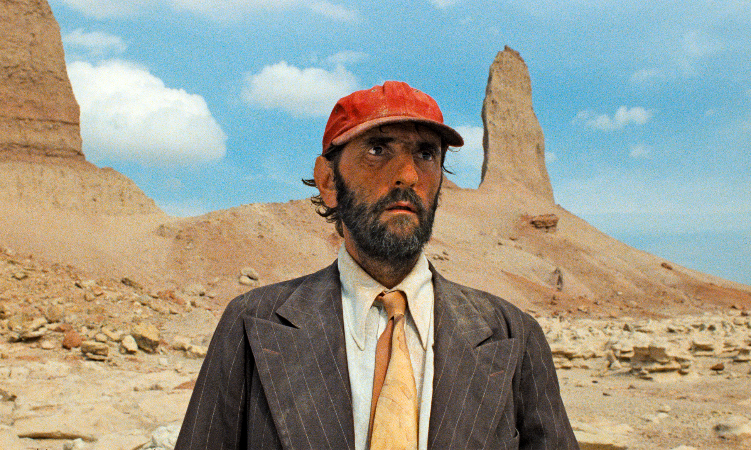 Plan iconique du film Paris, Texas de Wim Wenders. Un homme barbu, le regard perdu, porte une casquette rouge usée, une chemise blanche, une cravate beige et une veste à rayures marron. Il se tient debout dans un paysage désertique aride, au sol rocailleux, avec des formations rocheuses en arrière-plan sous un ciel bleu parsemé de nuages. L’homme semble désorienté et solitaire, incarnant la quête d’identité et de rédemption au cœur du film.