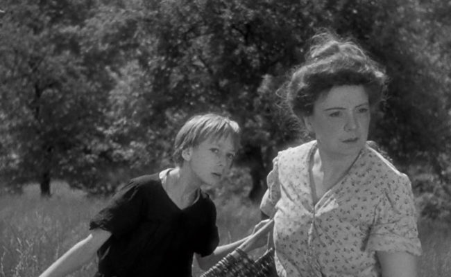Image en noir et blanc tirée du film Poil de Carotte. Au premier plan, une femme d’une trentaine d’années, coiffée d’un chignon, tient un grand panier en osier et regarde vers la droite, l’air inquiet. Derrière elle, le jeune garçon, vêtu d’un vêtement sombre, se penche légèrement en avant en regardant dans la même direction. La scène se déroule en extérieur, dans un champ bordé d’arbres, sous une lumière naturelle.