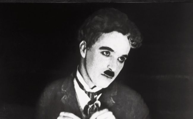 Charlot, personnage emblématique incarné par Charlie Chaplin, exécute la célèbre danse des petits pains, tenant deux fourchettes plantées dans des petits pains qu’il fait danser comme des marionnettes. Scène culte du film muet "La ruée vers l'or" en noir et blanc.