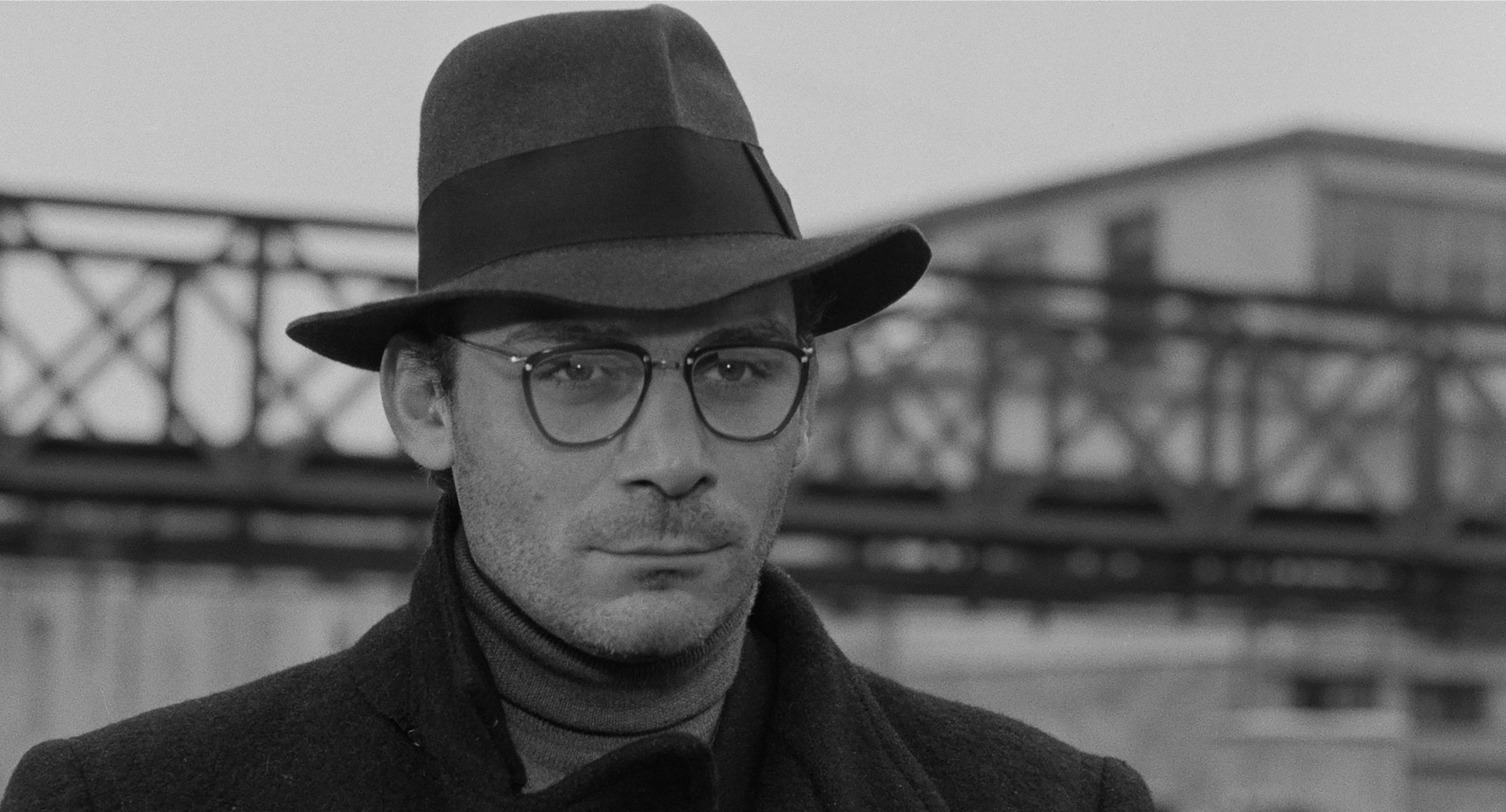 Un plan serré sur le visage du protagoniste en noir et blanc. Le personnage, impassible sous son feutre, incarne le visage humain du résistant, dans toute sa gravité et sa solitude. Le flou de l’arrière-plan isole davantage cette figure.