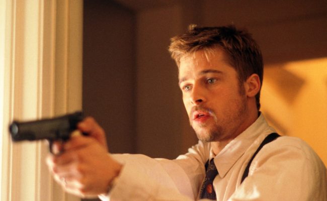 Un homme (Brad Pitt, dans le rôle du détective David Mills) en chemise blanche, cravate et bretelles tient une arme de poing à deux mains, pointée vers l’avant, avec une expression tendue et concentrée sur le visage. La scène est éclairée par une lumière chaude et se déroule à l'intérieur d'un bâtiment.