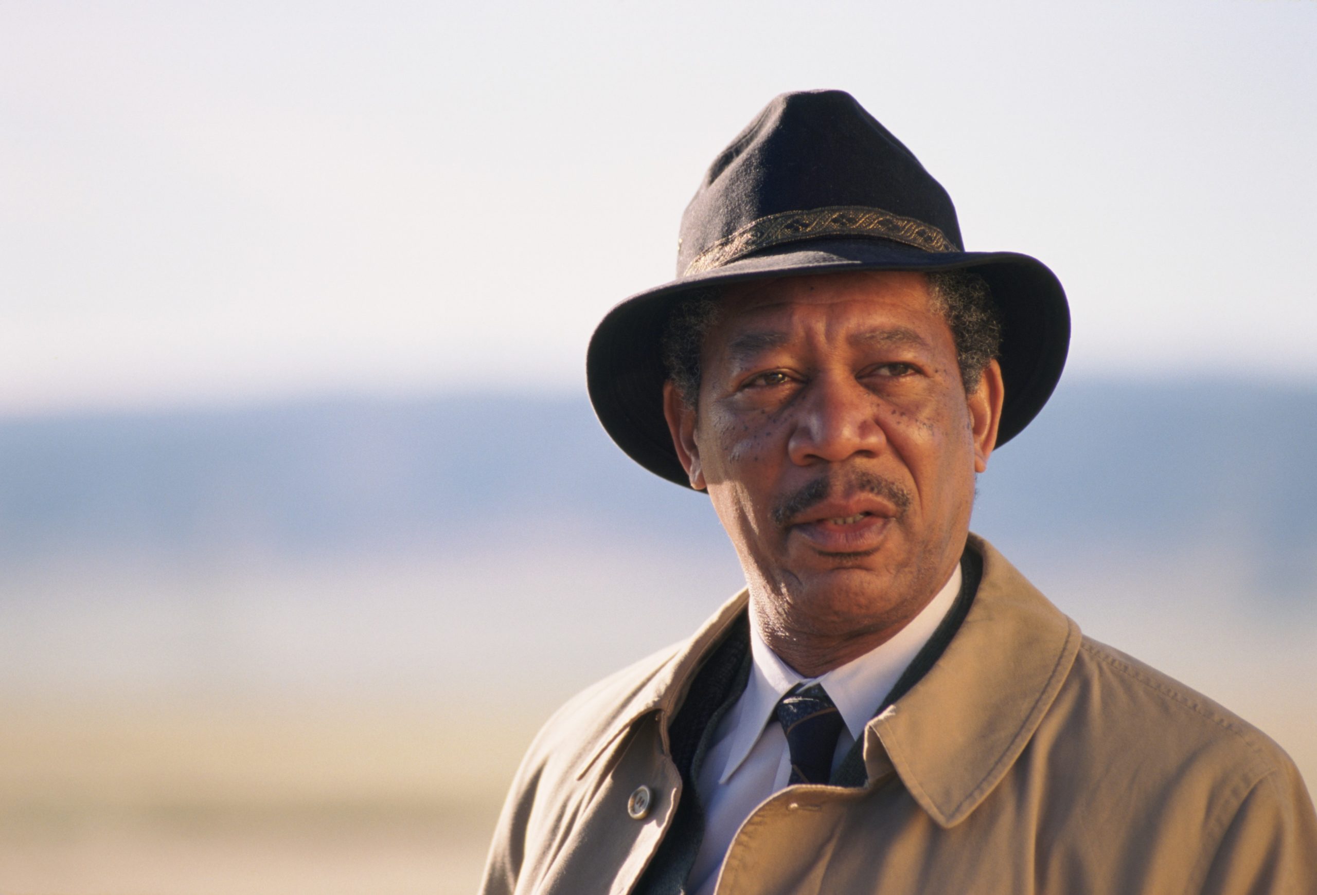 Plan rapproché du personnage de William Somerset, interprété par Morgan Freeman, dans le film Seven (1995). Il porte un chapeau sombre à motif doré, un imperméable beige et une cravate, et se tient à l'extérieur dans un décor de plaine lumineuse. Il regarde au loin avec une expression sérieuse et pensive, dans une lumière douce et naturelle.